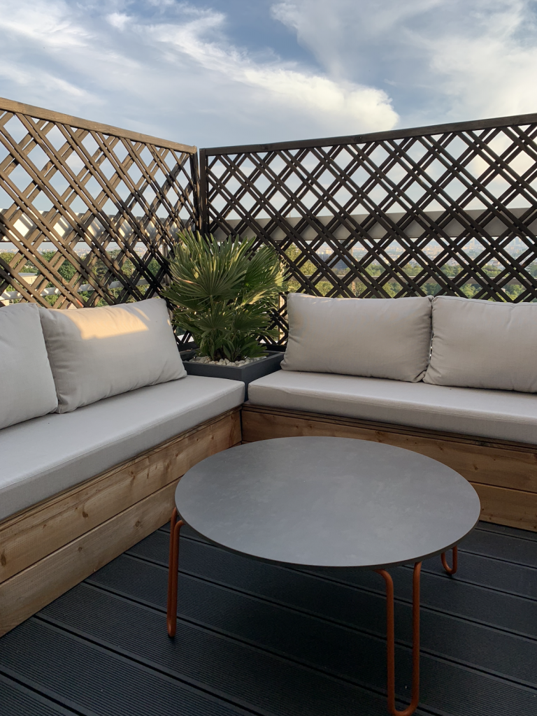 Aménagement de balcon avec banquette en bois sur mesure, coussins gris et brise-vue en losanges dans un appartement rénové par Home Feeling dans le Val d’Oise