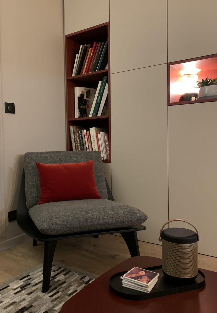 Fauteuil scandinave gris avec coussin rouge devant une bibliothèque encastrée sur mesure dans un appartement rénové par Home Feeling dans le Val d’Oise